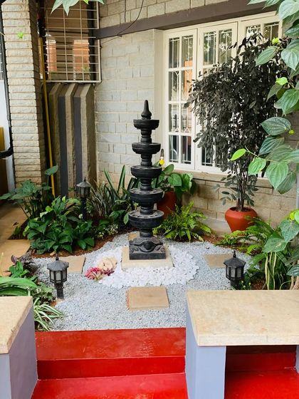 A wider view of the entrance garden featuring the Deepa Stambha. The mix of dark and light foliage, along with the traditional elements, creates a welcoming and culturally rich entryway.