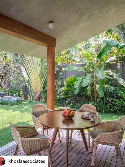 An outdoor dining set on a wooden deck, looking out onto a sunny lawn. The space is surrounded by tropical plants like banana trees, creating a perfect setting for a meal in a garden paradise.