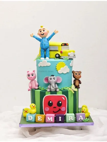 A two-tier Cocomelon cake for Demira's birthday, featuring JJ and the yellow school bus on top of a vibrant, character-filled design.