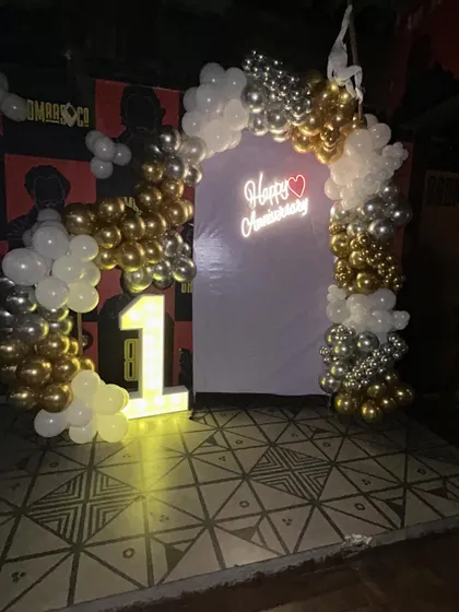 A classic anniversary setup at a commercial venue, combining a gold, silver, and white balloon arch with an illuminated marquee number and a "Happy Anniversary" neon sign for a truly celebratory feel.