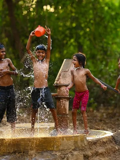 Four children gathered around the handpump, each engaged in the playful act of splashing water. The image showcases the teamwork and fun of a classic summer pastime.