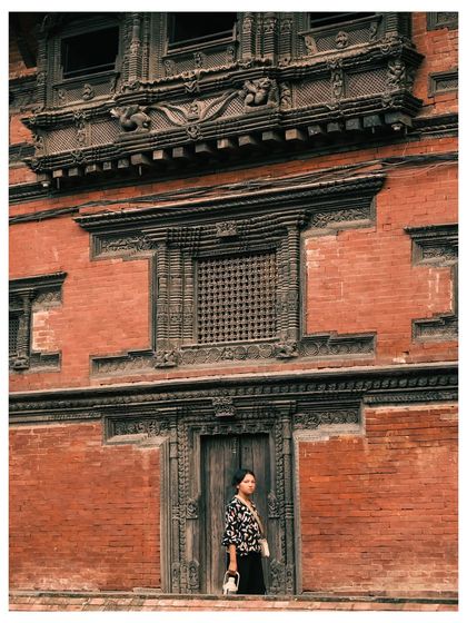 A woman stands in the doorway of an ancient building in Kathmandu, surrounded by intricate brickwork and carved wooden windows. The shot blends portraiture with architectural documentation.