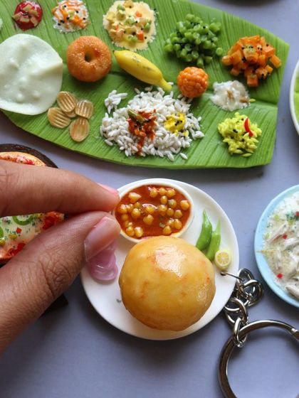 A close-up of a chole bhature keychain next to a South Indian banana leaf meal.