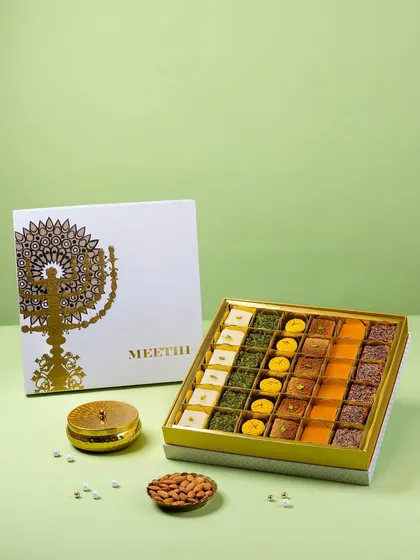 An open box of assorted Indian sweets for Meethi India. The composition includes the branded box lid and almonds to showcase the product's context and quality.
