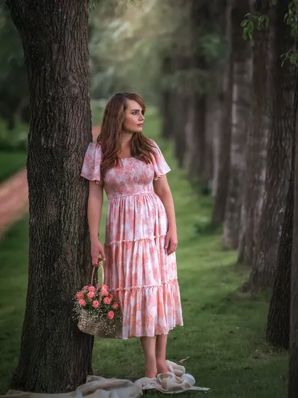 A dreamy outdoor portrait with a vintage feel. The woman stands barefoot by a line of trees, holding a basket of flowers. The soft focus and warm light give this image a romantic, almost ethereal quality, like a scene from a classic novel.