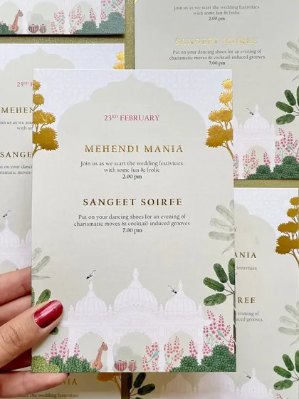 Holding one of the event cards from the Udaipur suite. This card for the Mehendi and Sangeet features a serene illustration of the palace grounds, inviting guests to an evening of festivities.