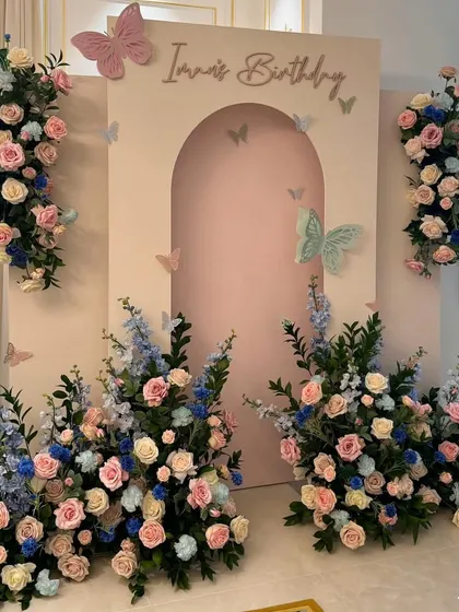 An elegant floral birthday setup with a pastel pink arched backdrop, surrounded by beautiful arrangements of pink, blue, and white flowers for a soft, dreamy look.