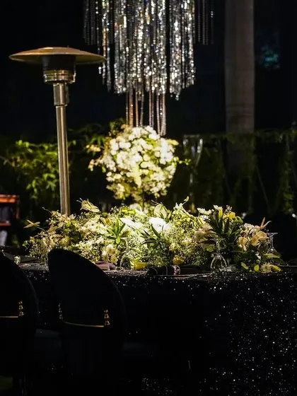 A table at a cocktail party, featuring a black sequined tablecloth, a centerpiece of white and green florals, and a modern tassel chandelier.