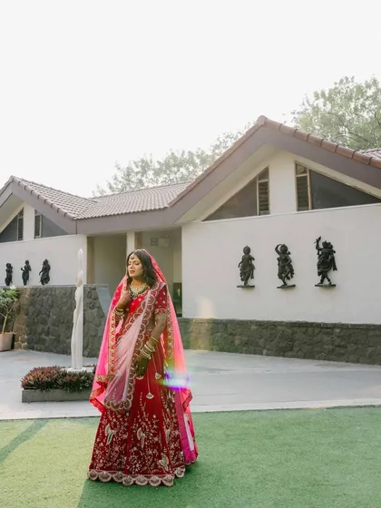 A full-length bridal portrait of Sakshi. The modern architecture of the venue provides a clean and interesting backdrop for her vibrant red and pink lehenga.