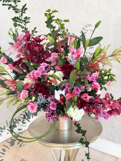 A congratulatory arrangement in a symphony of pinks and burgundies. The mix of cymbidium orchids, roses, and lilies, with a trailing vine of ivy, creates a look of abundant celebration.