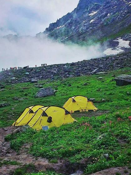 Tents pitched on a vibrant green meadow, with misty clouds rolling through the mountain pass. This is a scene from a group trek, where the journey is as beautiful as the destination.