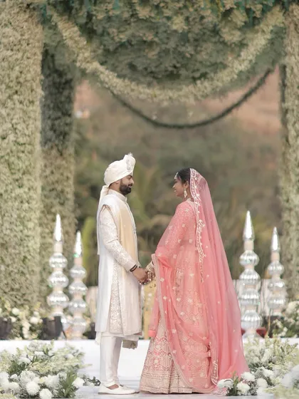 The couple holds hands after the varmala ceremony, sharing a quiet moment of connection. The soft lighting and beautiful floral decor create a romantic and intimate atmosphere.