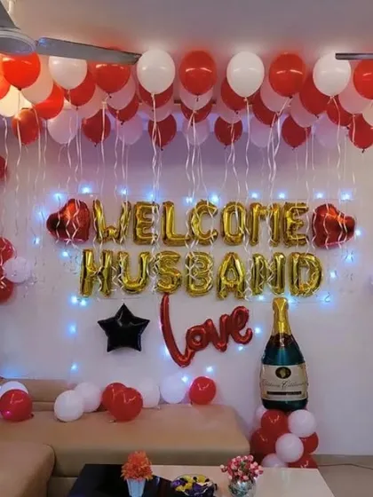 A special "Welcome Husband" decoration for a living room space. This setup uses red and white balloons, foil letters, and a large champagne bottle balloon to create a fun and loving welcome party atmosphere.