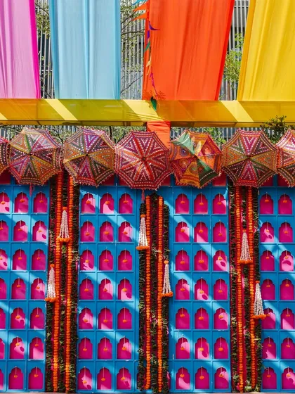 A vibrant Rajasthani-mela style photo wall, with a blue jharokha-style panel, marigold garlands, and colorful umbrellas.