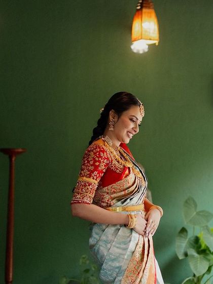 A bride in a light blue and red saree, her posture elegant and poised, stands on the green-walled verandah.