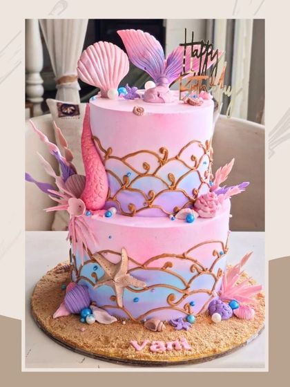 A stunning two-tier mermaid cake with an ombre pink and purple wave design, golden accents, and intricate seashell and coral details.
