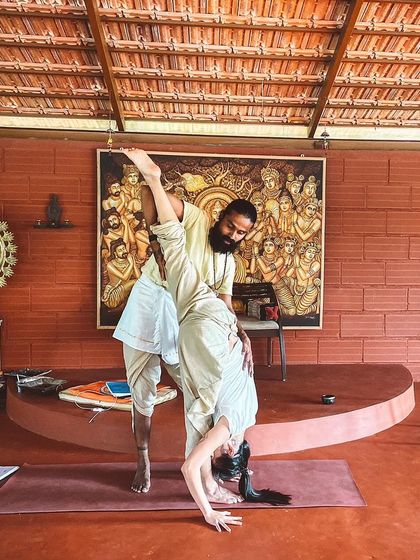 I assist a student in a standing forward bend with a leg lift, a posture that requires both strength and surrender. My role is to provide stability so the student can explore their edge with confidence.