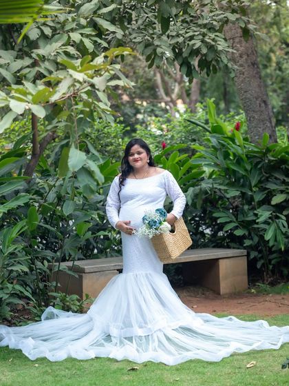 A serene portrait of a mom-to-be in a white gown, standing in a park and holding a basket with blue flowers.