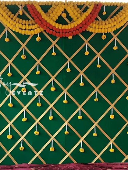 A traditional backdrop design with a green fabric base and a gold ribbon lattice pattern. The decor is framed with yellow marigolds and accented with small hanging floral bunches.