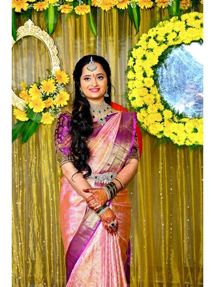 A full shot against a festive yellow floral backdrop. The makeup is kept soft and natural with a pop of pink on the lips to match her saree.