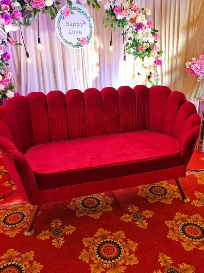 A close-up of the luxurious red velvet shell-style sofa. This statement piece of furniture adds a touch of vintage glamour and vibrant color to any romantic event or photo booth area.