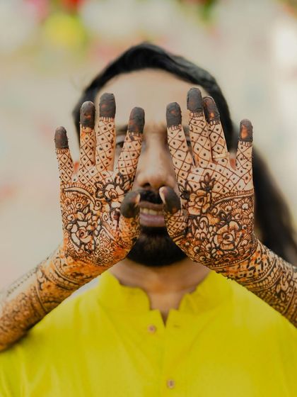 A playful Mehendi moment where the groom peeks through his hands, covered in intricate henna designs. It’s a fun, candid shot that captures the lighter side of wedding traditions.