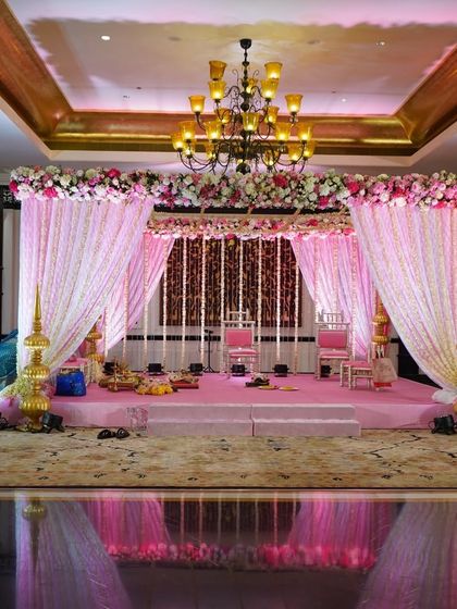 A beautiful pastel pink mandap setup. The stage is framed by drapes and florals in soft pink hues, with traditional golden lamps, creating a serene and romantic space for a wedding ceremony.