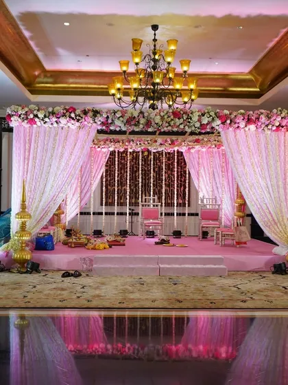 A beautiful mandap setup for a pastel-themed wedding, with a floral canopy, soft pink drapes, and traditional golden pillars, all reflected on a mirrored floor.