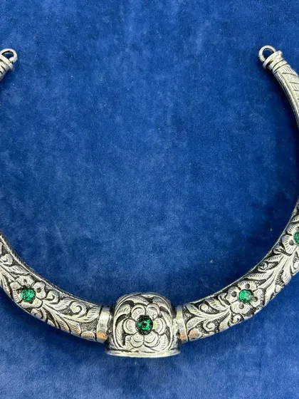 A premium pure silver hasli, or rigid choker. This piece is hand-carved with a beautiful floral pattern and studded with small green stones, offering a classic and elegant look.