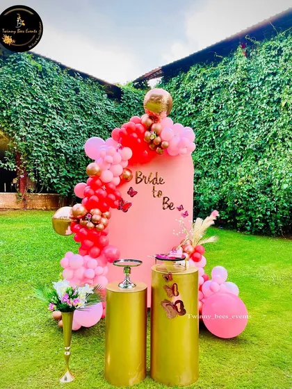 This outdoor "Bride to be" setup features a pink backdrop decorated with a cascade of red and pink balloons and gold butterfly accents. The gold pedestals are ready for the cake and desserts.