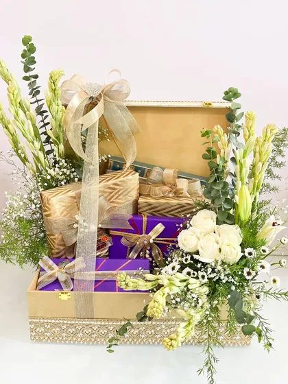 A luxurious gift trunk for a roka, filled with beautifully wrapped presents in purple and gold, and adorned with an arrangement of fresh white flowers.