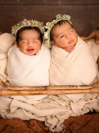 Two newborn twins, swaddled in white and wearing floral wreaths, are pictured sleeping together in a rustic wooden crib. This heartwarming shot captures the special bond between twins from their very first days.