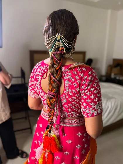 A fun and festive braid for a Mehendi ceremony. The hair is woven with colorful orange and pink threads and decorated with vibrant tassels, perfectly matching the bride's playful outfit.
