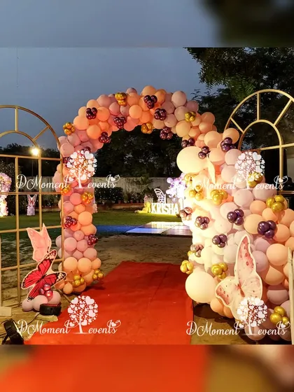 A beautiful golden-arched gateway for an outdoor butterfly-themed party. The entrance is adorned with a cascading balloon arrangement in peach and purple tones, with a red carpet leading guests into the celebration.