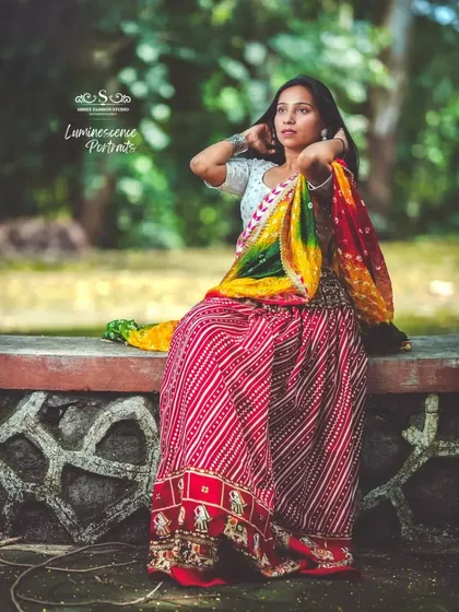 A full-length portrait of a woman in a colorful Bandhani lehenga, seated gracefully on a stone ledge in a park.