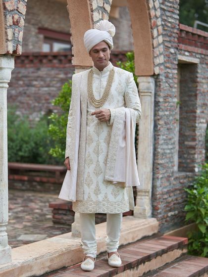This ivory sherwani, with its subtle gold embroidery, is captured against a traditional backdrop, emphasizing its timeless appeal. The complete look is one of pure elegance and heritage.