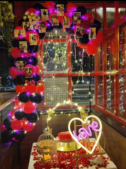 This balcony setup for a couple features a balloon arch decorated with their photos, a table covered in rose petals, and a neon 'Love' sign, creating a personalized and romantic corner.