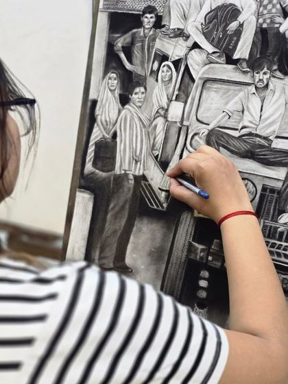 A close-up shot of the artist's hand, showing the careful application of charcoal to bring out the details in the clothing of the figures in her drawing.