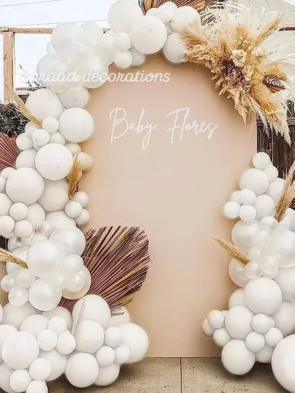 A chic boho-themed baby shower backdrop. This design features an all-white balloon garland accented with dried pampas grass and palm leaves against a neutral-toned arched wall, creating a trendy and modern look.