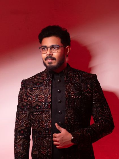 A stylish groom-to-be looking off-camera, captured against a dramatic red background that highlights his sharp, modern sherwani.