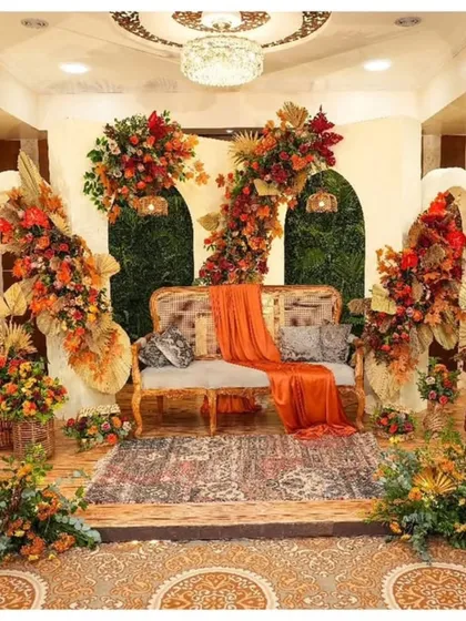 A rich, rustic-themed stage setup perfect for a Haldi or Mehendi ceremony. I used a warm, autumnal color palette with orange and red flowers, dried palm leaves, and pampas grass, all centered around a comfortable cane sofa to create a cozy and photogenic space.