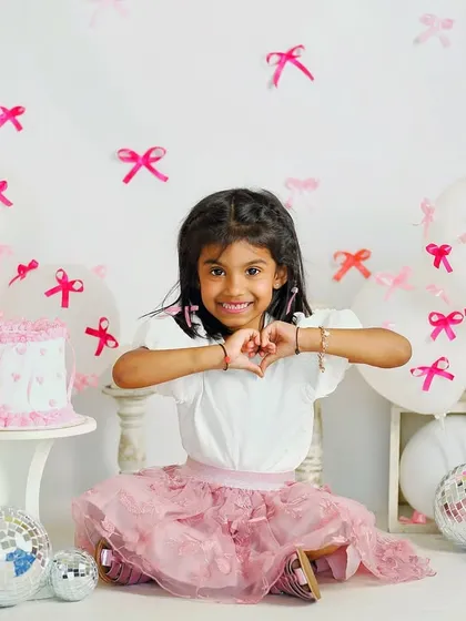 Celebrating a fifth birthday with a pink and white bow-themed party setup. The birthday girl is making a heart sign, surrounded by balloons and disco balls.