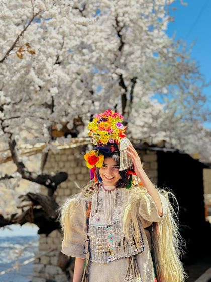 A smiling woman from the Brokpa tribe in Ladakh, her colorful headdress a beautiful contrast to the white blossoms behind her. Her joy is a reflection of the beauty of her homeland.