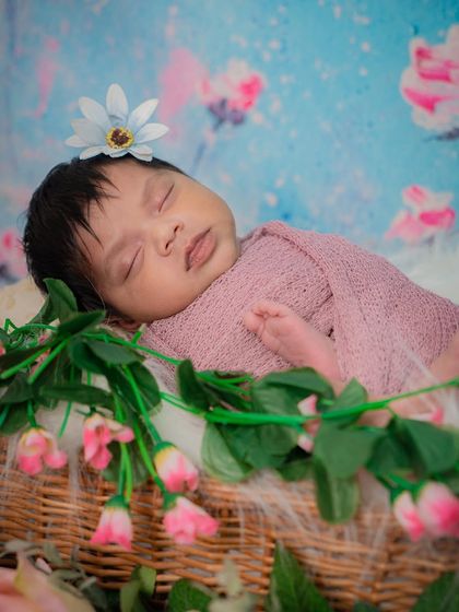 A beautiful newborn girl sleeping in a basket adorned with pink flowers and greenery. The floral backdrop adds a dreamy, garden-like feel to the portrait.