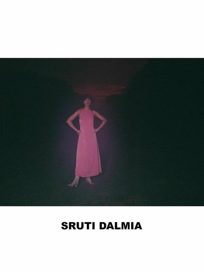A dramatic, high-contrast image from the Sruti Dalmia shoot. The model is styled in a simple pink dress, set against a dark landscape with a fiery sunset sky, creating a powerful and evocative silhouette.