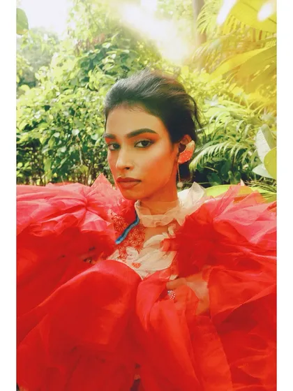 A vibrant, high-color image from the 'Roots' editorial. The bright red tulle and bold orange makeup create a powerful contrast with the lush green background, symbolizing a blossoming of identity.