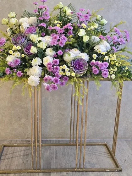 A beautiful floral arrangement on a modern gold stand. This piece combines purple and white flowers with ornamental cabbage for a unique texture and color palette.