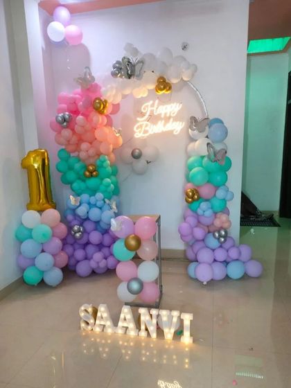 A whimsical pastel rainbow theme for a first birthday. This home decoration includes colorful balloon pillars, a 'Happy Birthday' neon sign, and light-up letters spelling the birthday girl's name.