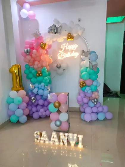 A whimsical pastel rainbow theme for a first birthday. This home decoration includes colorful balloon pillars, a 'Happy Birthday' neon sign, and light-up letters spelling the birthday girl's name.