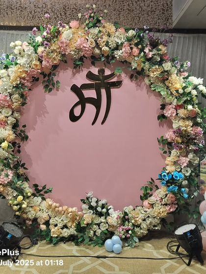 A beautiful baby shower backdrop with a large floral ring surrounding the 'Maa' (Mother) symbol in Hindi. The setup is flanked by cute Krishna and Radha cutouts on balloon pillars.